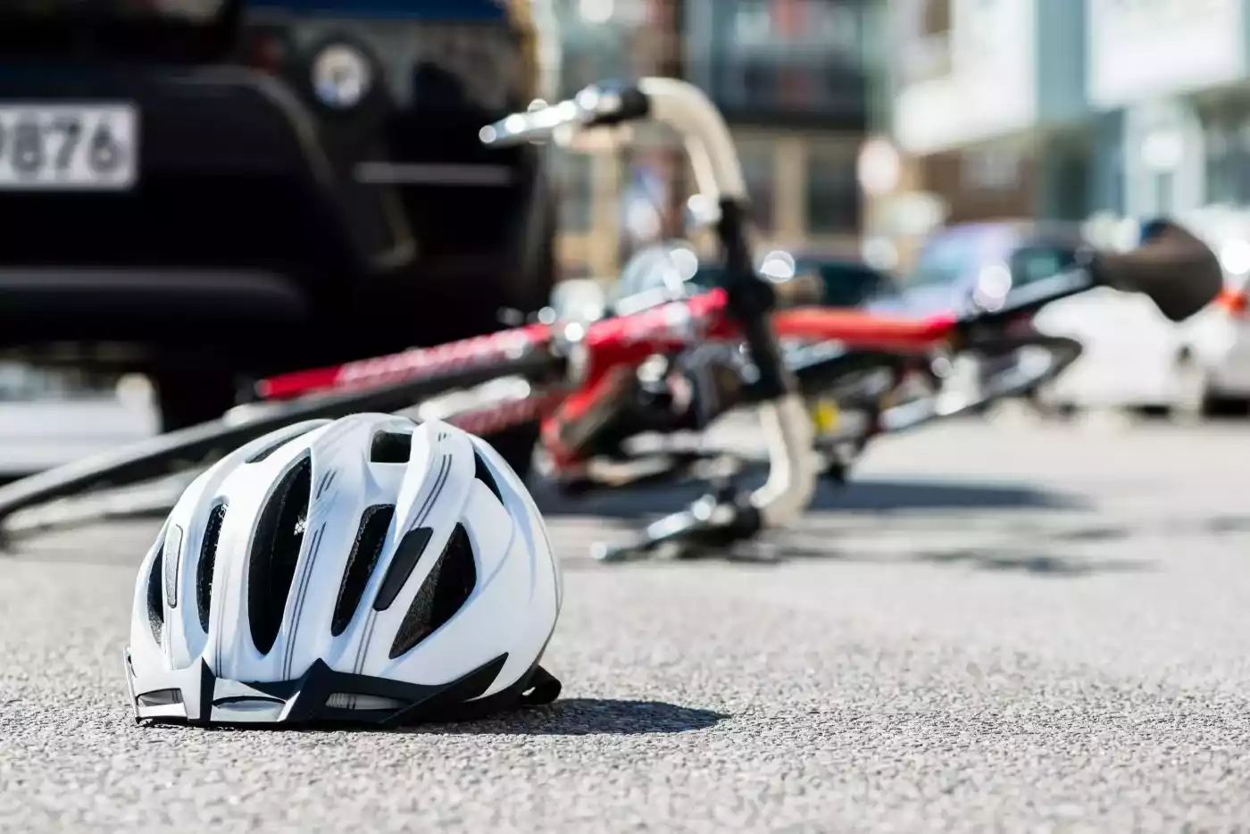 A white and black bicycle helmet lies on the asphalt in the foreground, with a fallen red bicycle and a car blurred in the background of a street scene.