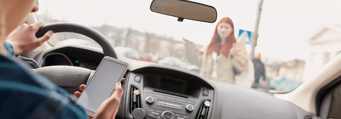 A driver looking down at a smartphone while a pedestrian stands in front of the car windshield.