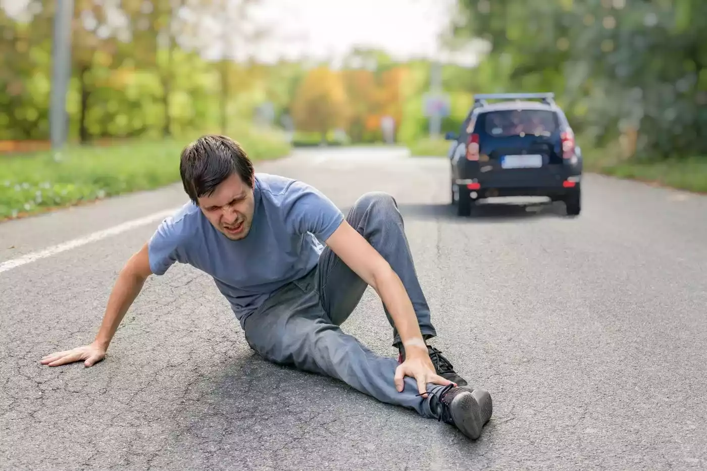 A man sitting in agony on an asphalt road holding his leg after a hit-and-run car accident.