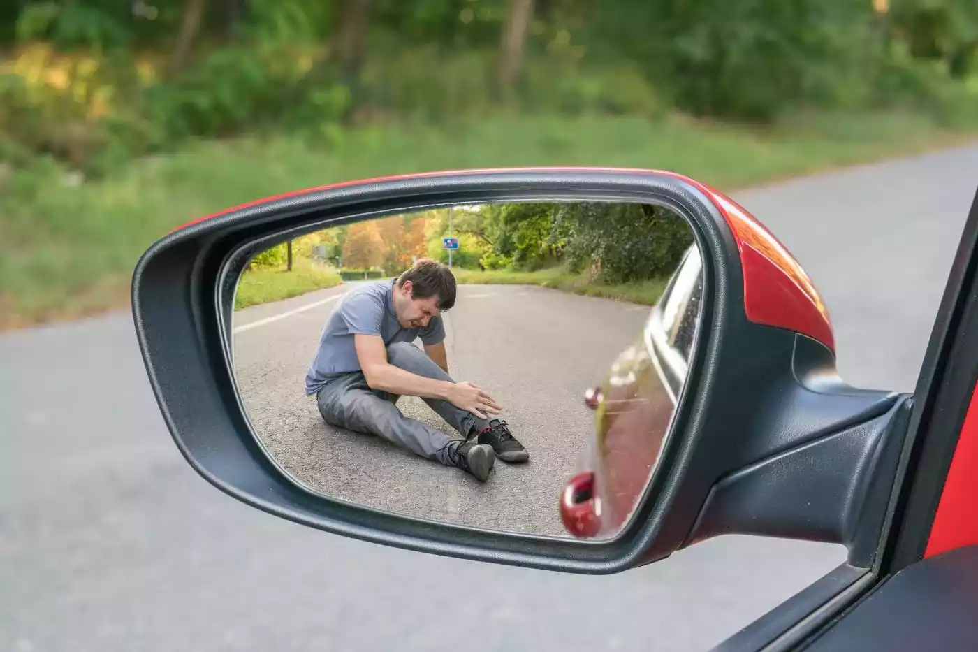 A man clutching his ankle on the road, visible through a car's side-view mirror.