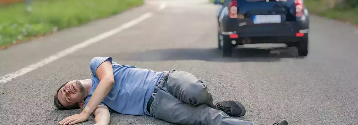 A person lying on the asphalt in front of a dark vehicle.