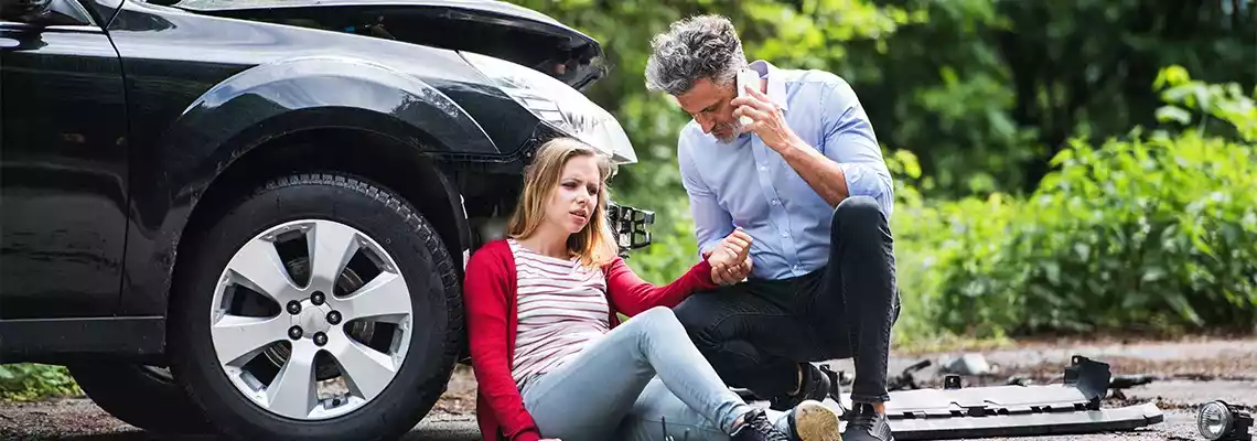 A man on the phone tends to an injured woman sitting next to a damaged black car after an accident.