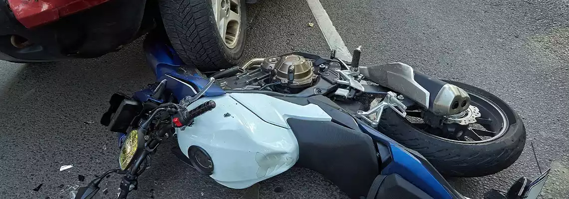 A motorcycle lies crashed on its side with its front end pinned underneath a car's tire.