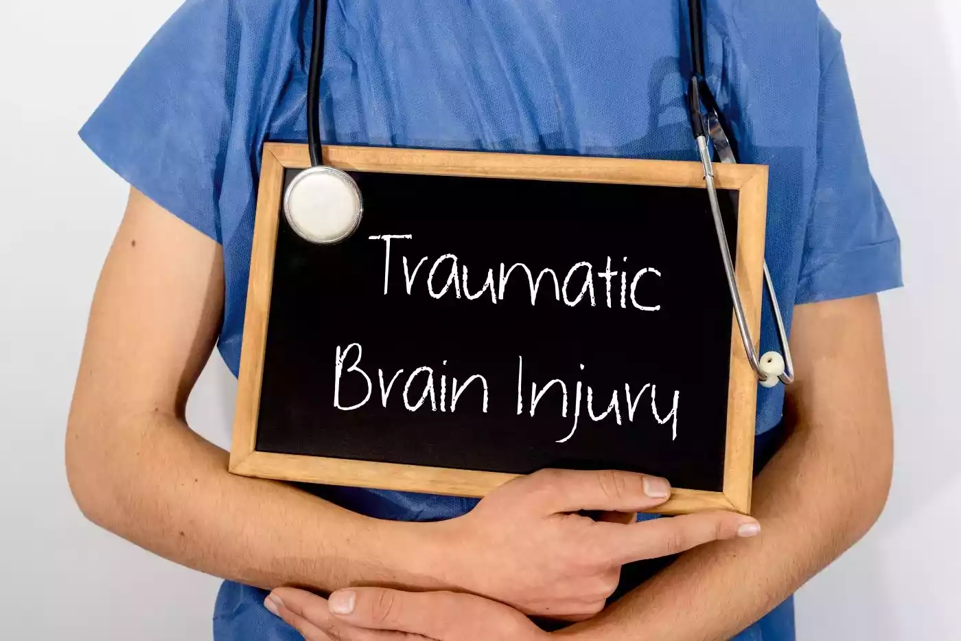 A person in blue scrubs holding a small chalkboard that reads 'Traumatic Brain Injury'