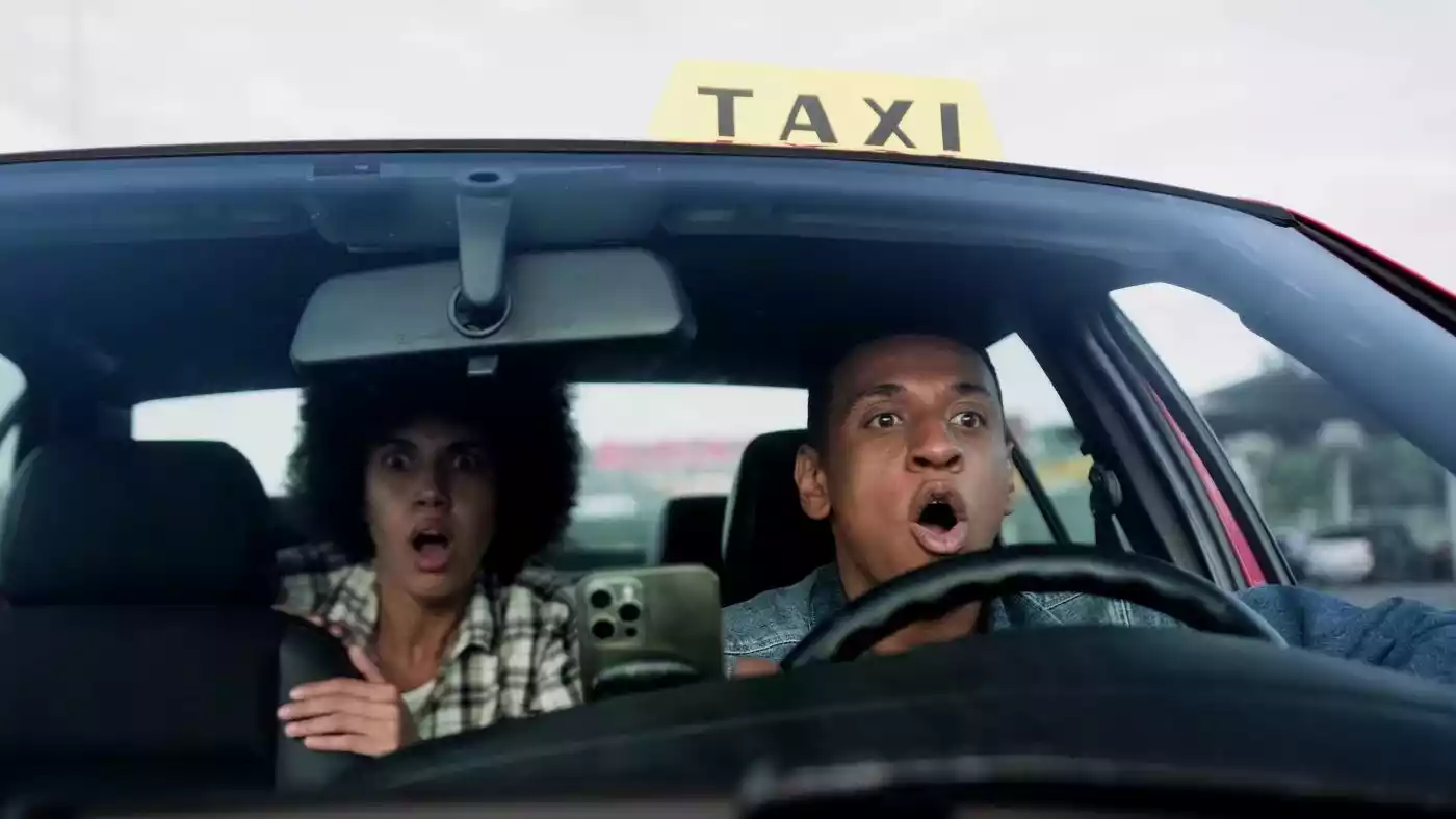 A taxi driver and a passenger look straight ahead with wide eyes and open mouths in shocked panic.