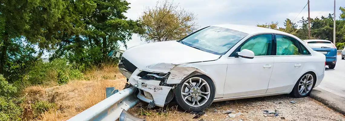 A white sedan with severe front-end damage crashed into a metal guardrail on the side of a road.