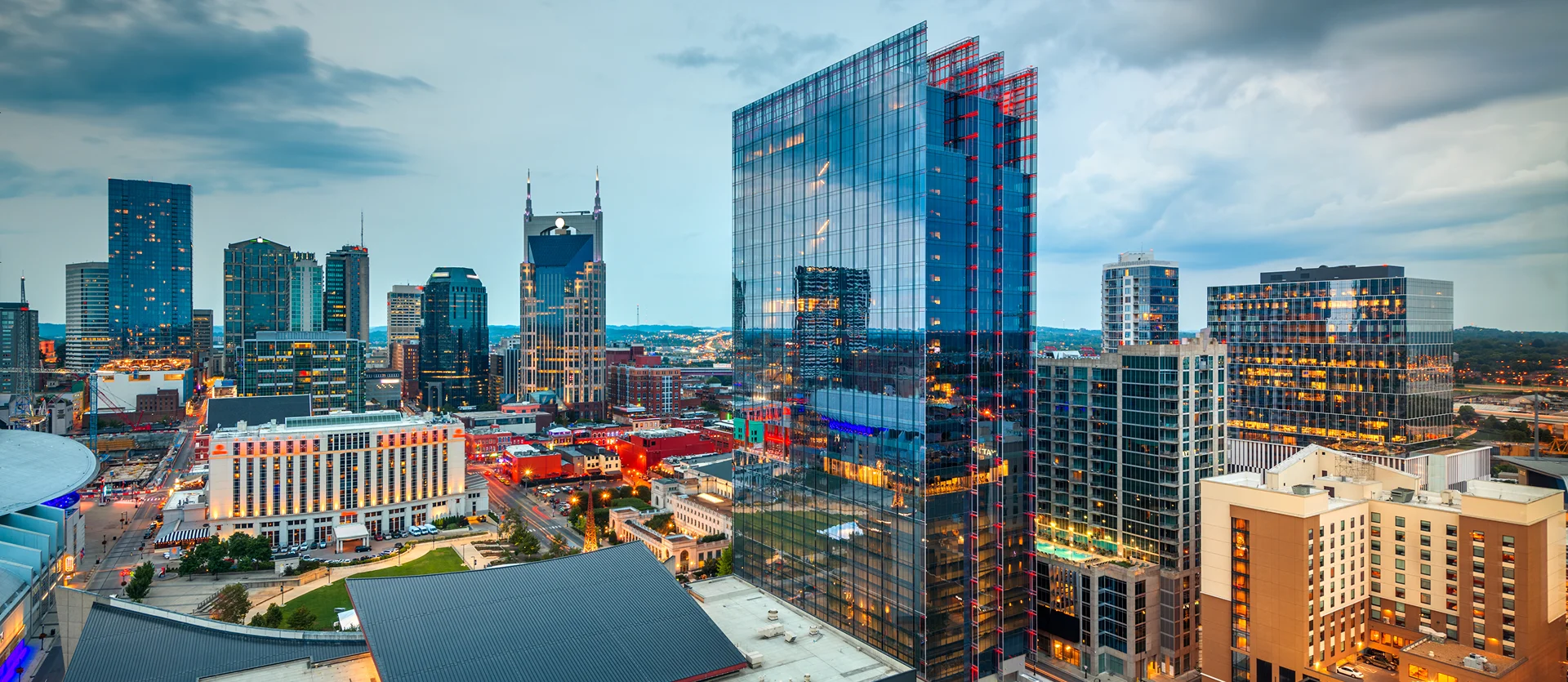 Panoramic view of a city skyline at dusk featuring modern glass skyscrapers and illuminated buildings under a cloudy sky.