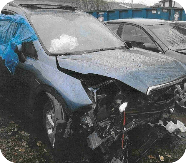 Damaged blue car with missing front bumper and windshield covered by blue tarp, parked next to another car.
