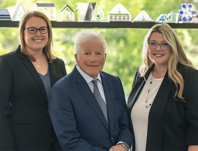 Three attorneys standing together in an office, smiling, with awards and a window behind them.
